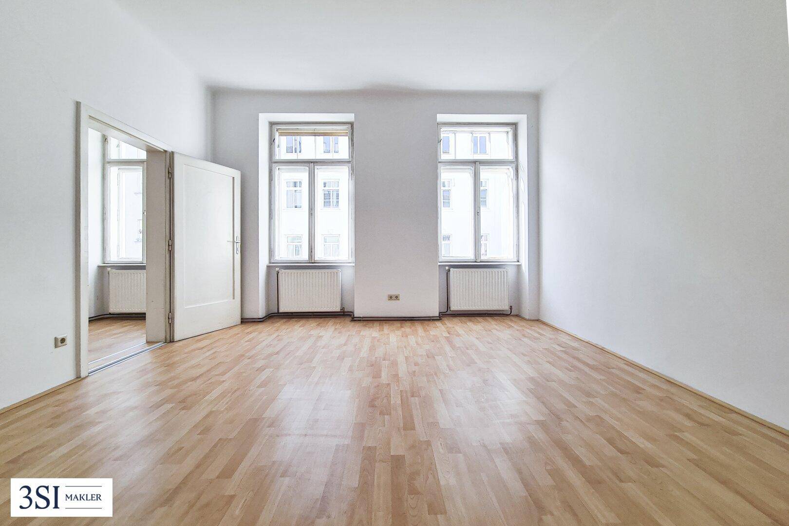 Leeres Wohnzimmer mit Holzboden, weißen Wänden und zwei großen Fenstern.