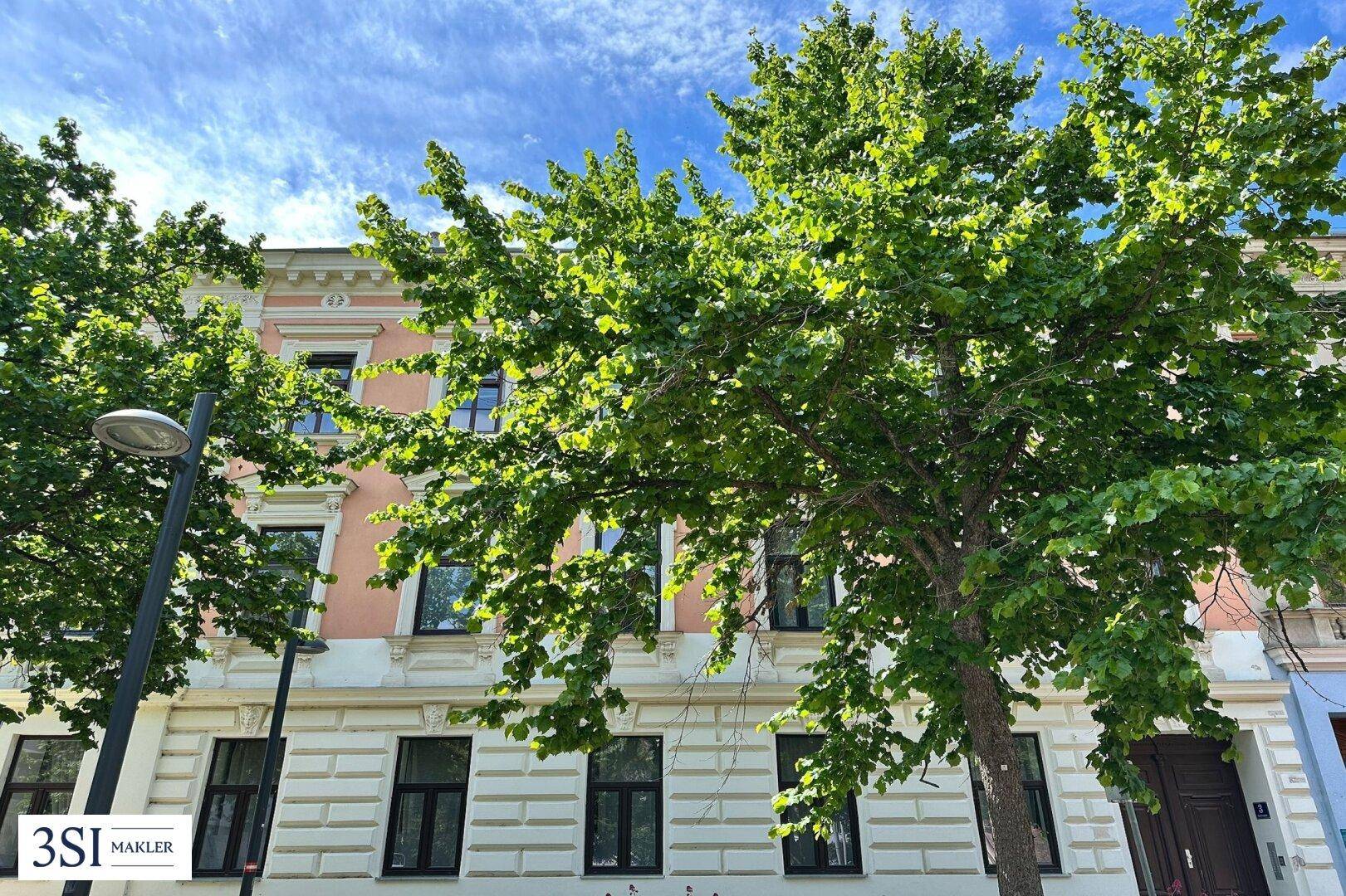Historische Fassade eines Wohngebäudes mit grünen Bäumen im Vordergrund unter blauem Himmel.