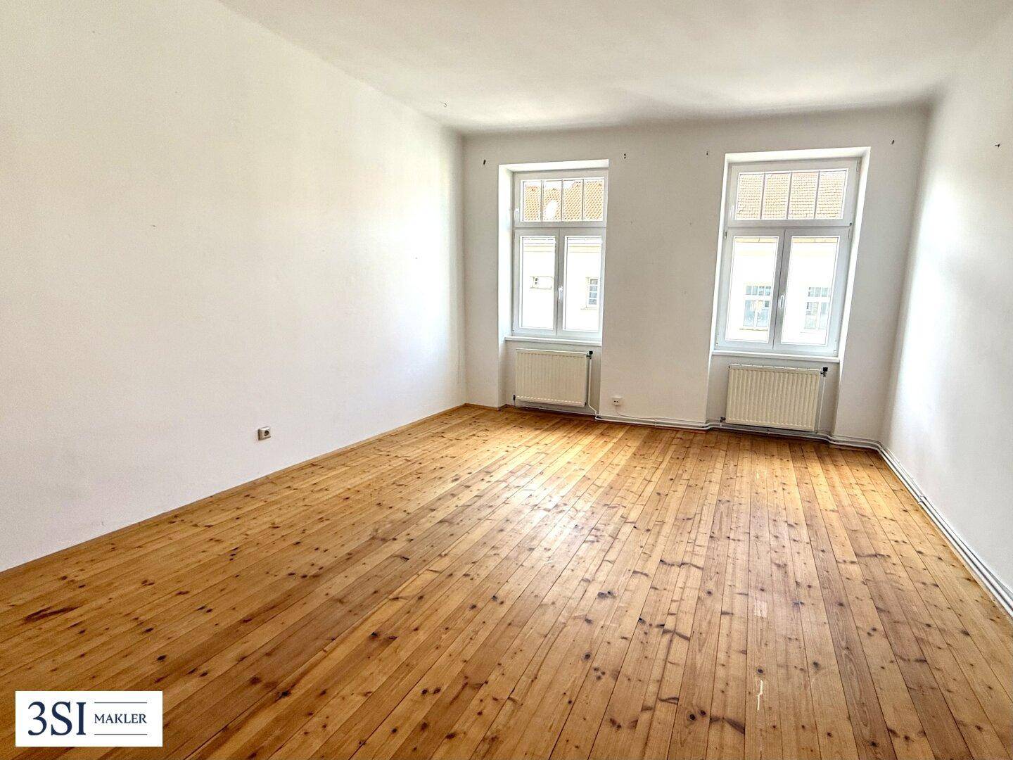 Leerer Raum mit Holzdielenboden, weißen Wänden und zwei großen Fenstern.