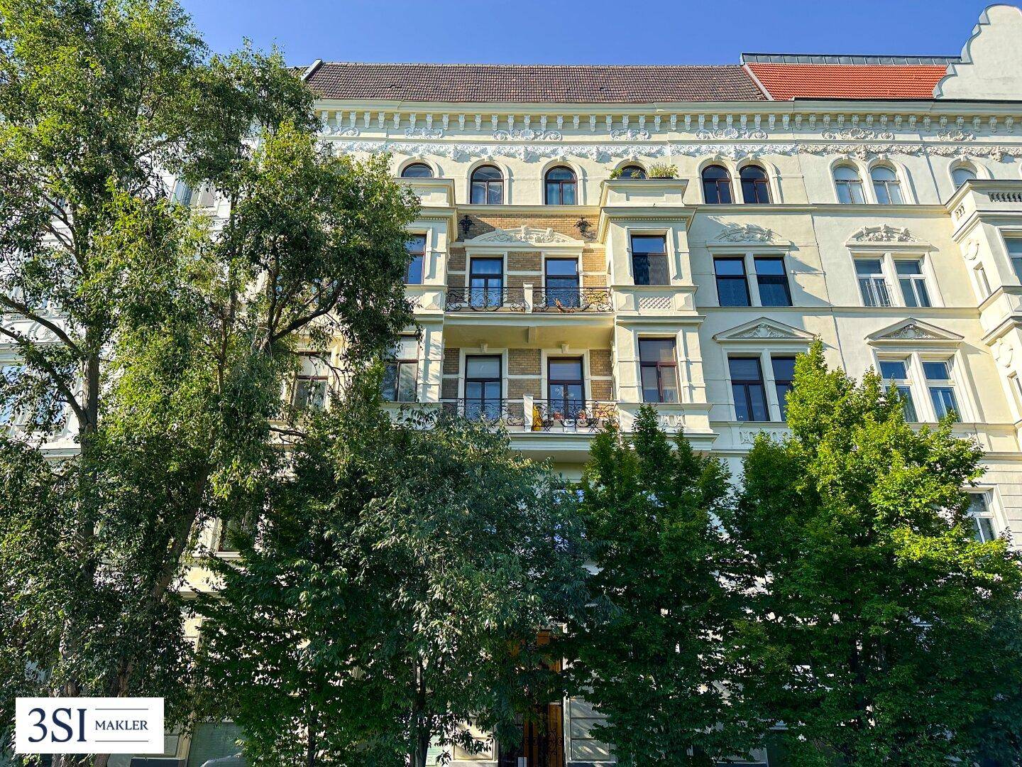 Historische Gebäudefassade mit mehreren Balkonen, großen Fenstern und umgeben von hohen Bäumen.
