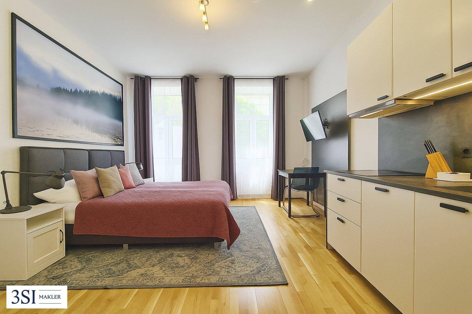 Helles Schlafzimmer mit Doppelbett, Holzboden und integrierter Küchenzeile.