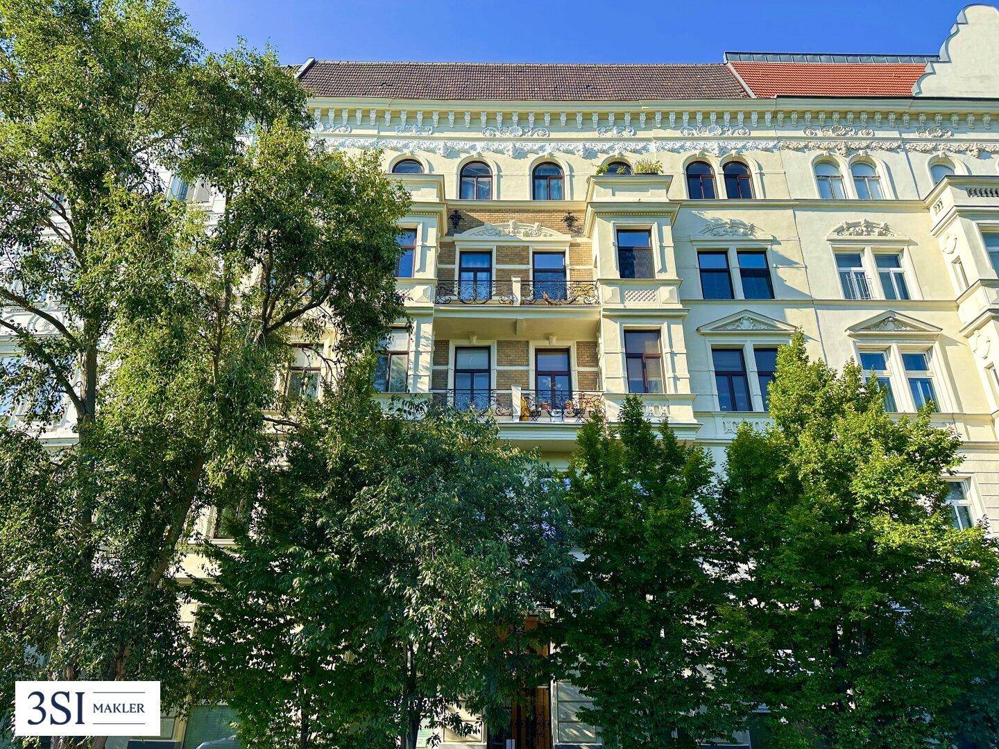 Historische Fassade eines Mehrfamilienhauses mit Balkonen und üppigem Baumbestand.