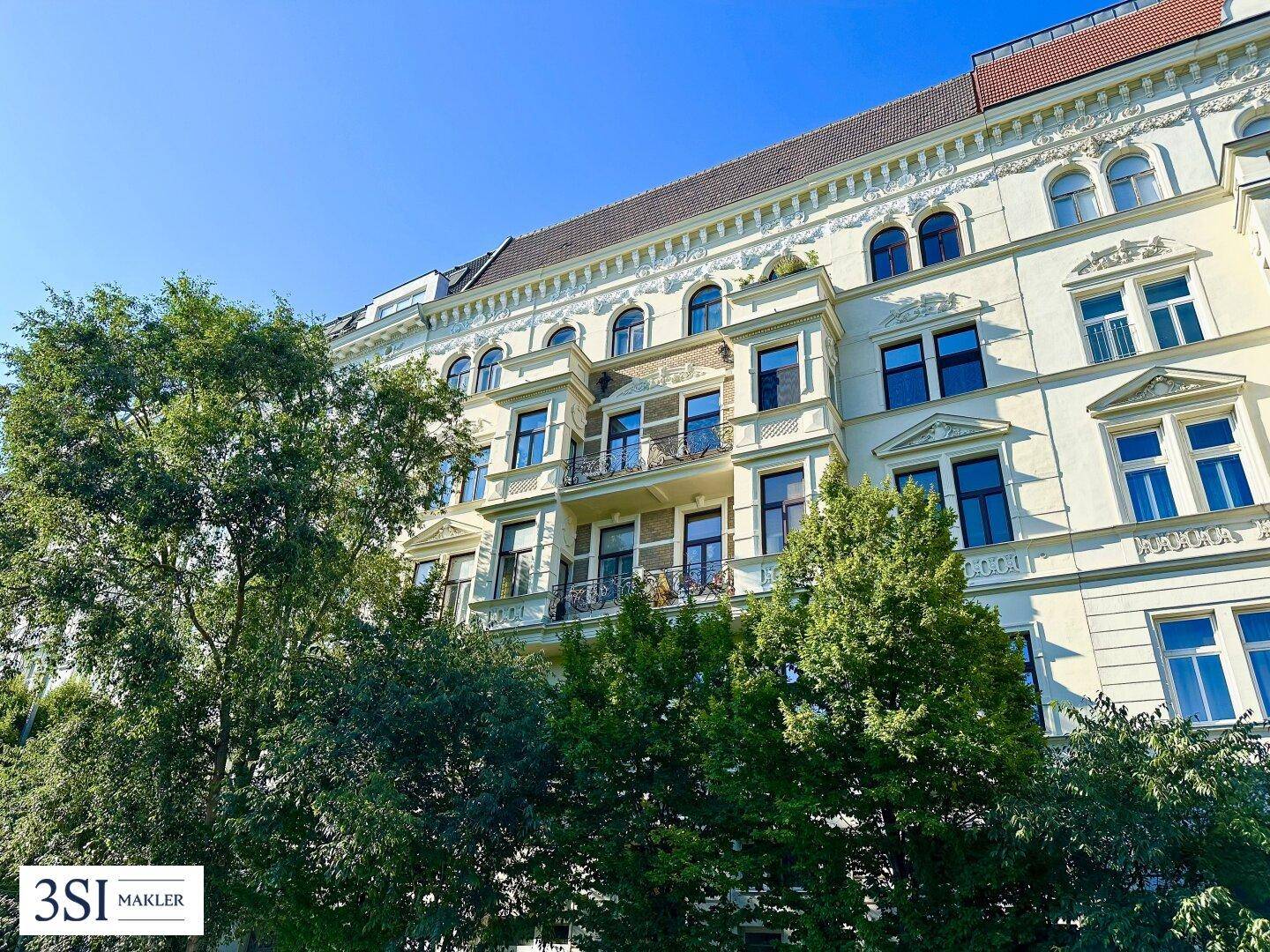 Historische Fassade eines Mehrfamilienhauses mit verzierten Balkonen und grüner Umgebung.