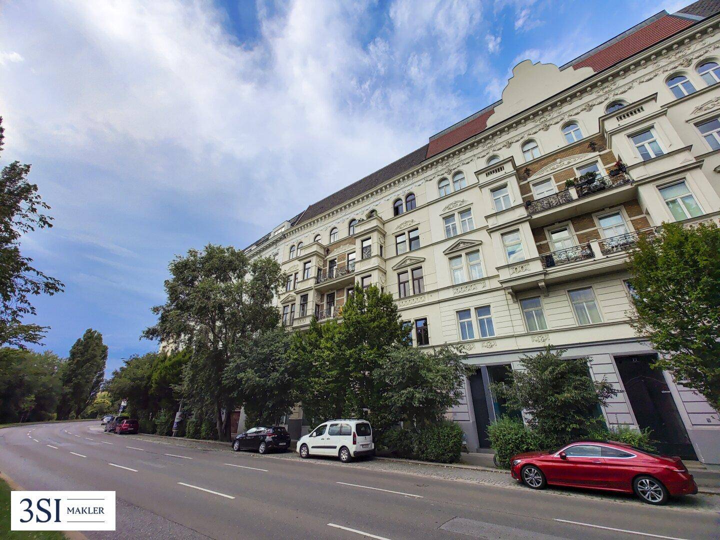 Gebäudeansicht von der Straße aus, mit parkenden Autos und Bäumen im Vordergrund.