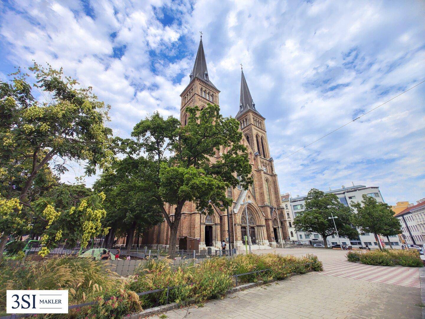 Historische Kirche mit zwei Türmen und Parkanlage unter bewölktem Himmel.