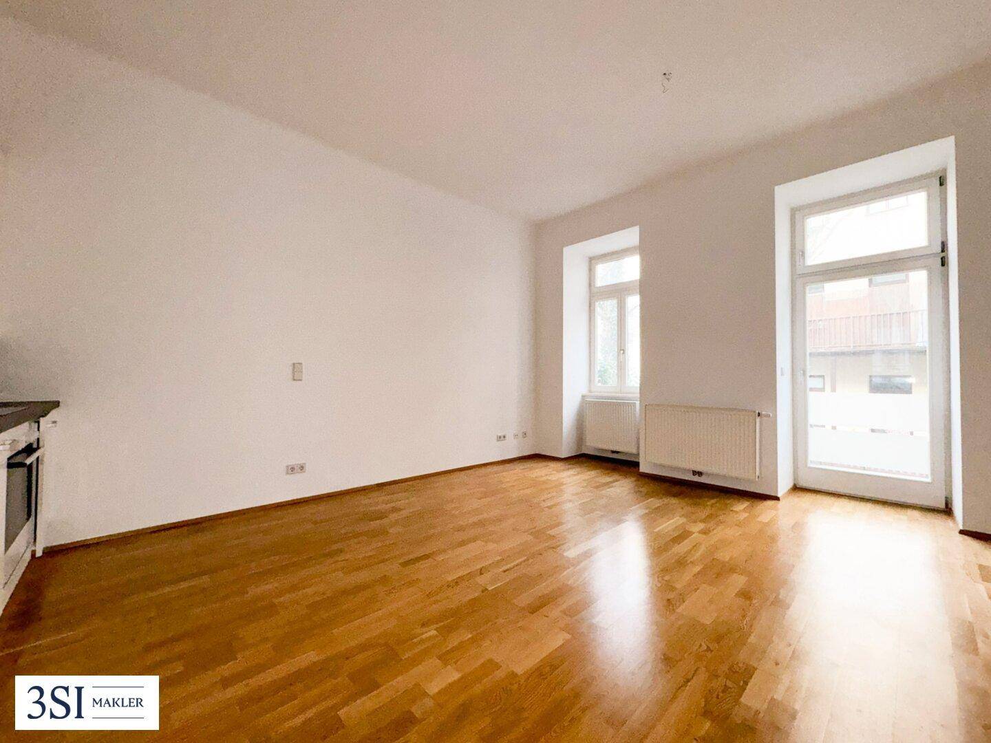 Geräumiger, leerer Wohnbereich mit Holzboden und viel Tageslicht durch große Fenster.