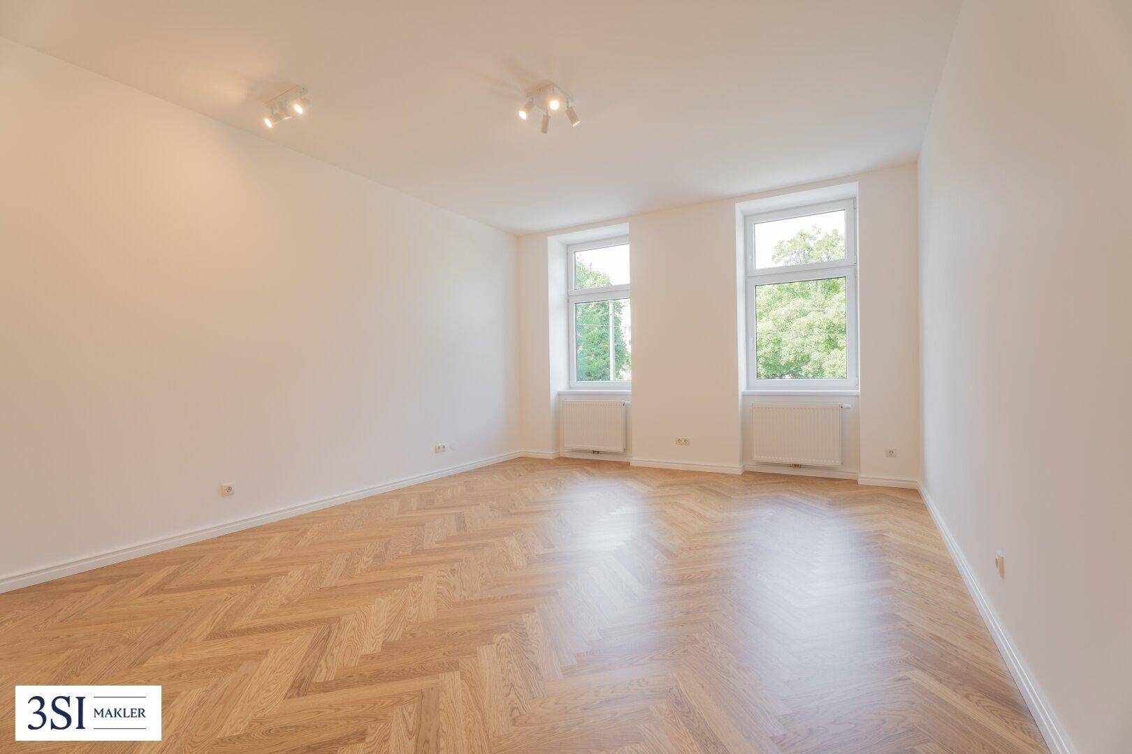 Geräumiges, helles Zimmer mit Fischgrätparkett und zwei großen Fenstern, bereit zur Einrichtung.