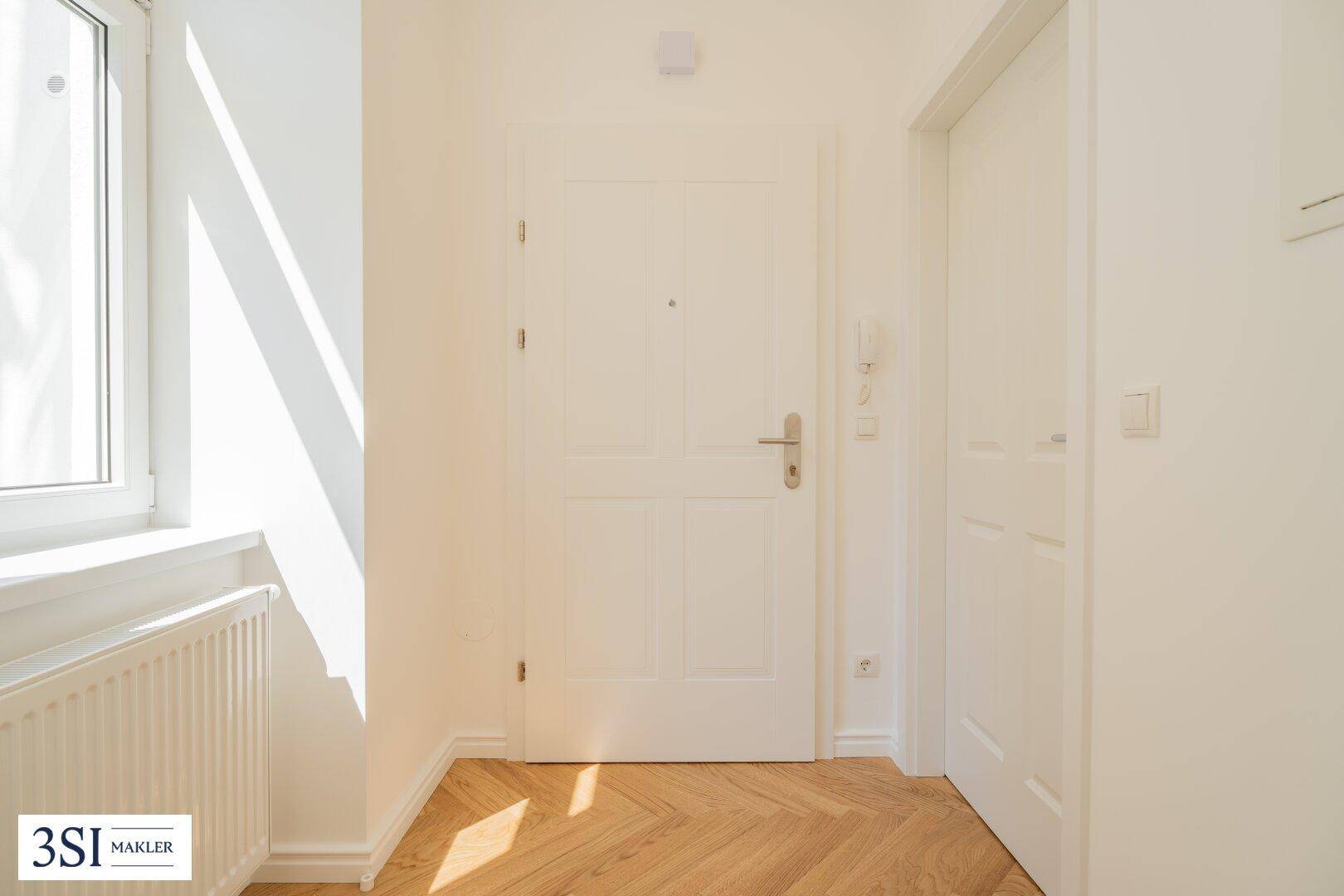 Eingangsbereich mit weißer Wohnungstür, Fischgrätparkett und einem Fenster, das Sonnenlicht hereinlässt.