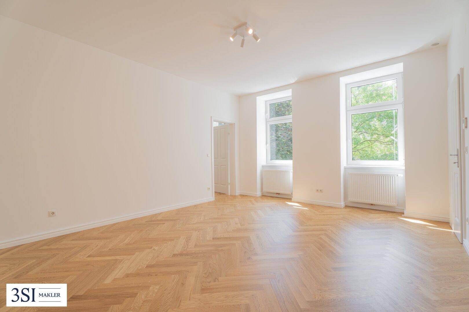 Geräumiges, leeres Wohnzimmer mit Fischgrätparkett und großen Fenstern, die viel Licht hereinlassen.