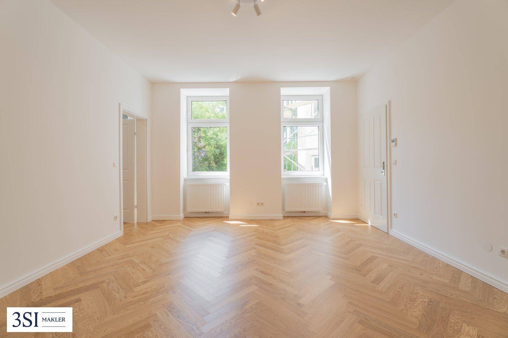 Helles, unmöbliertes Zimmer mit Fischgrätparkett und zwei großen Fenstern mit Blick ins Grüne.