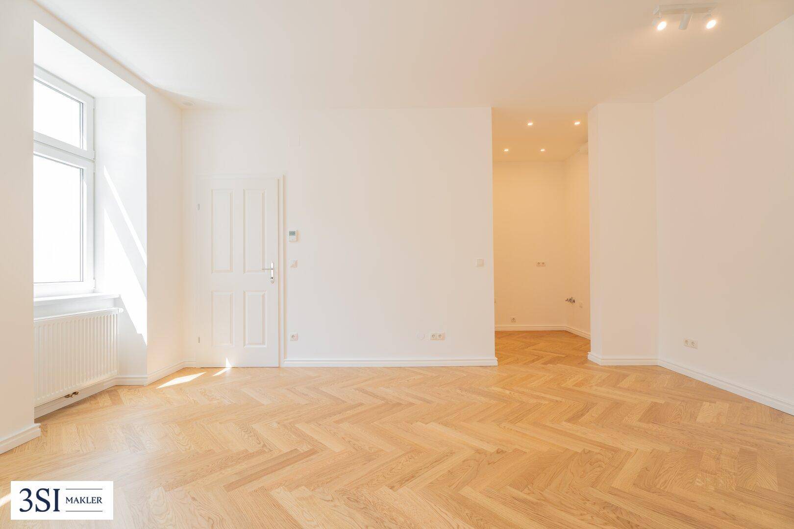 Heller, leerer Raum mit Fischgrätparkett und einem Fenster, das viel Sonnenlicht hereinlässt.
