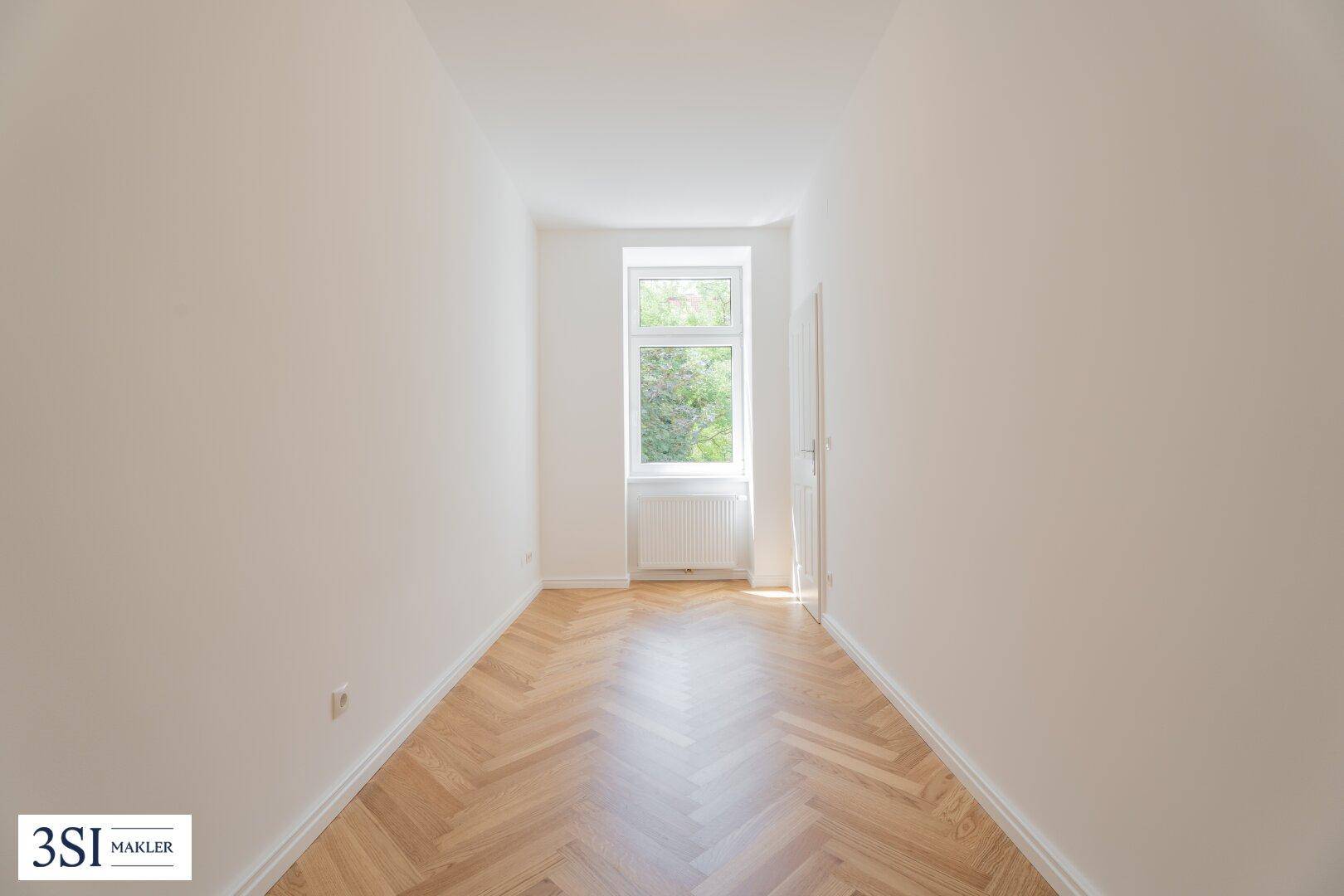 Langes, schmales Zimmer mit Fischgrätparkett und einem Fenster, das einen Blick ins Grüne bietet.