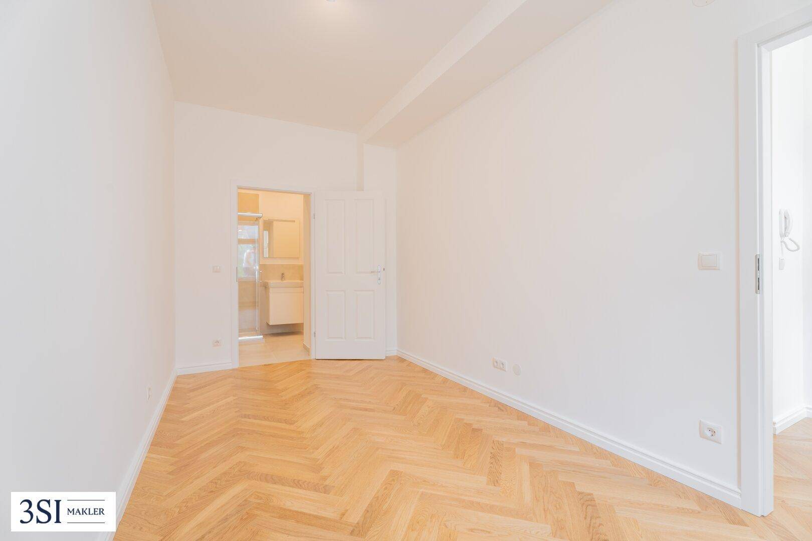 Heller Raum mit Fischgrätparkett und Blick in ein modernes Badezimmer.