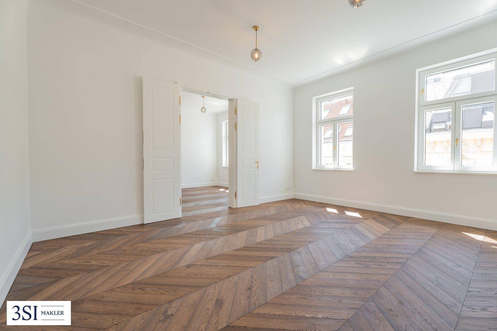 Geräumiges Zimmer mit Fischgrätparkett, hohen Decken und Flügeltüren zu einem weiteren Raum.