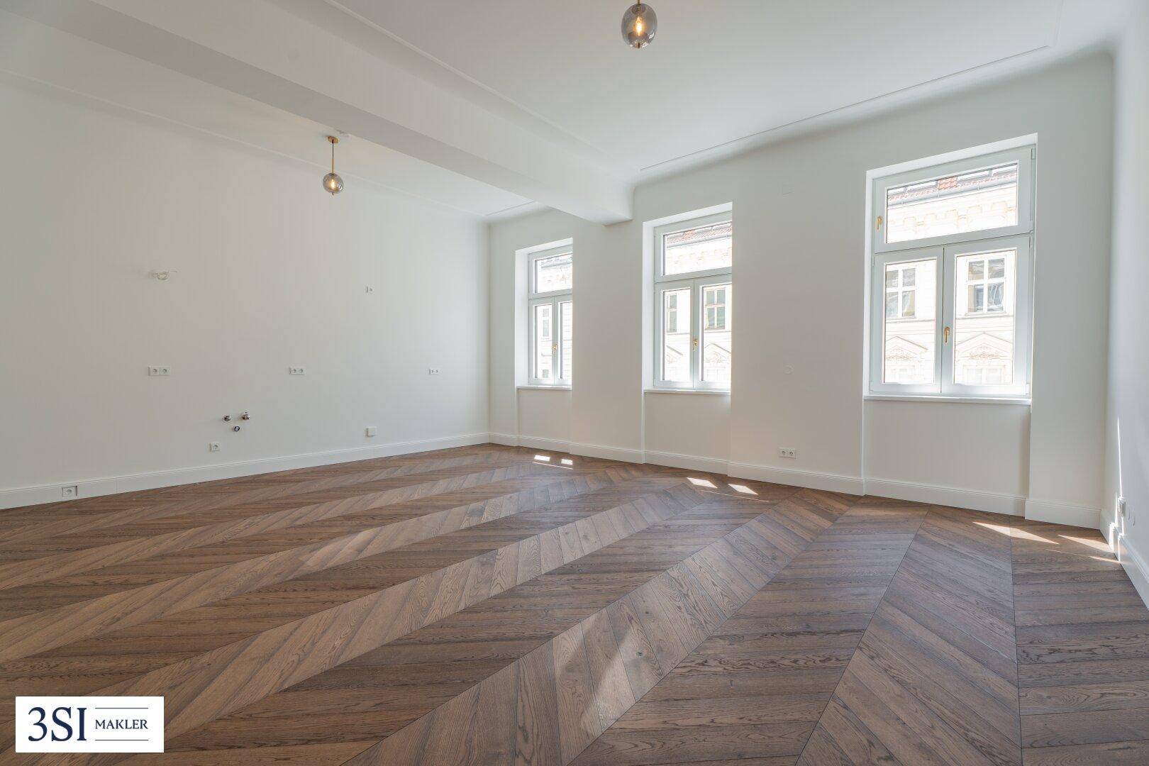 Großes, helles Zimmer mit Fischgrätparkett, hohen Decken und drei Fenstern.