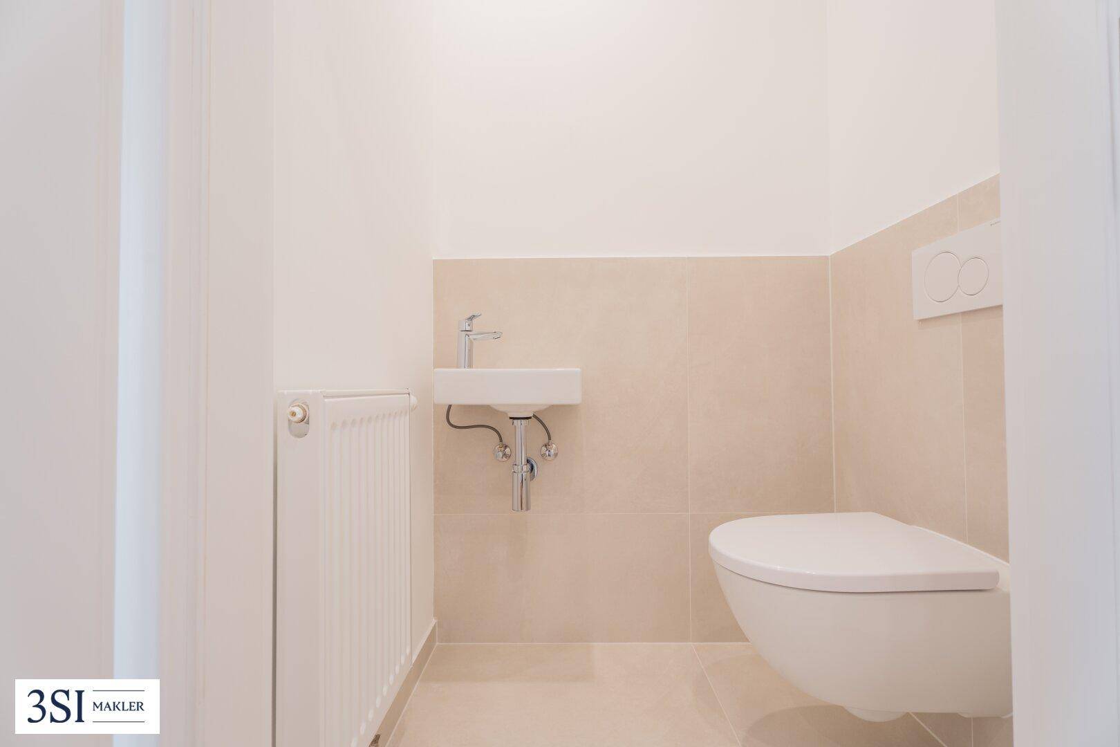 Separates WC mit wandhängender Toilette, kleinem Waschbecken und Heizkörper, in hellen Farben gehalten.