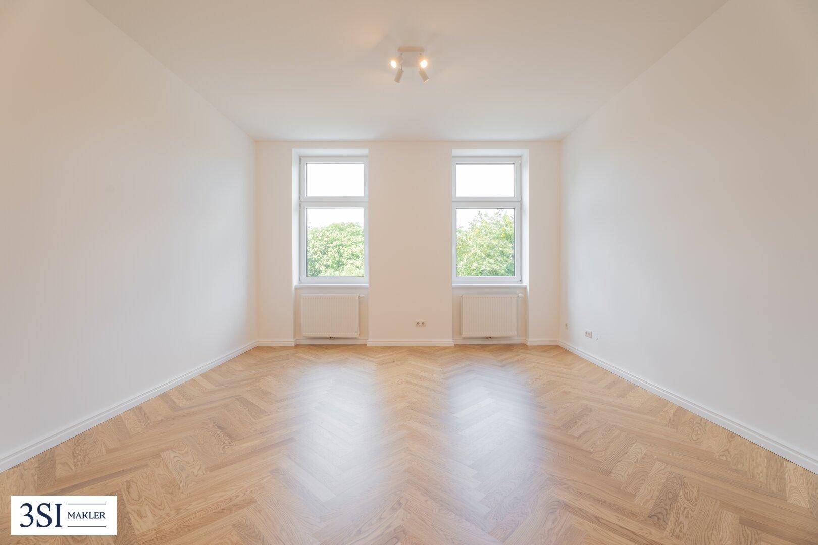 Geräumiges, leeres Zimmer mit Fischgrätparkett und zwei großen Fenstern, die viel Licht spenden.