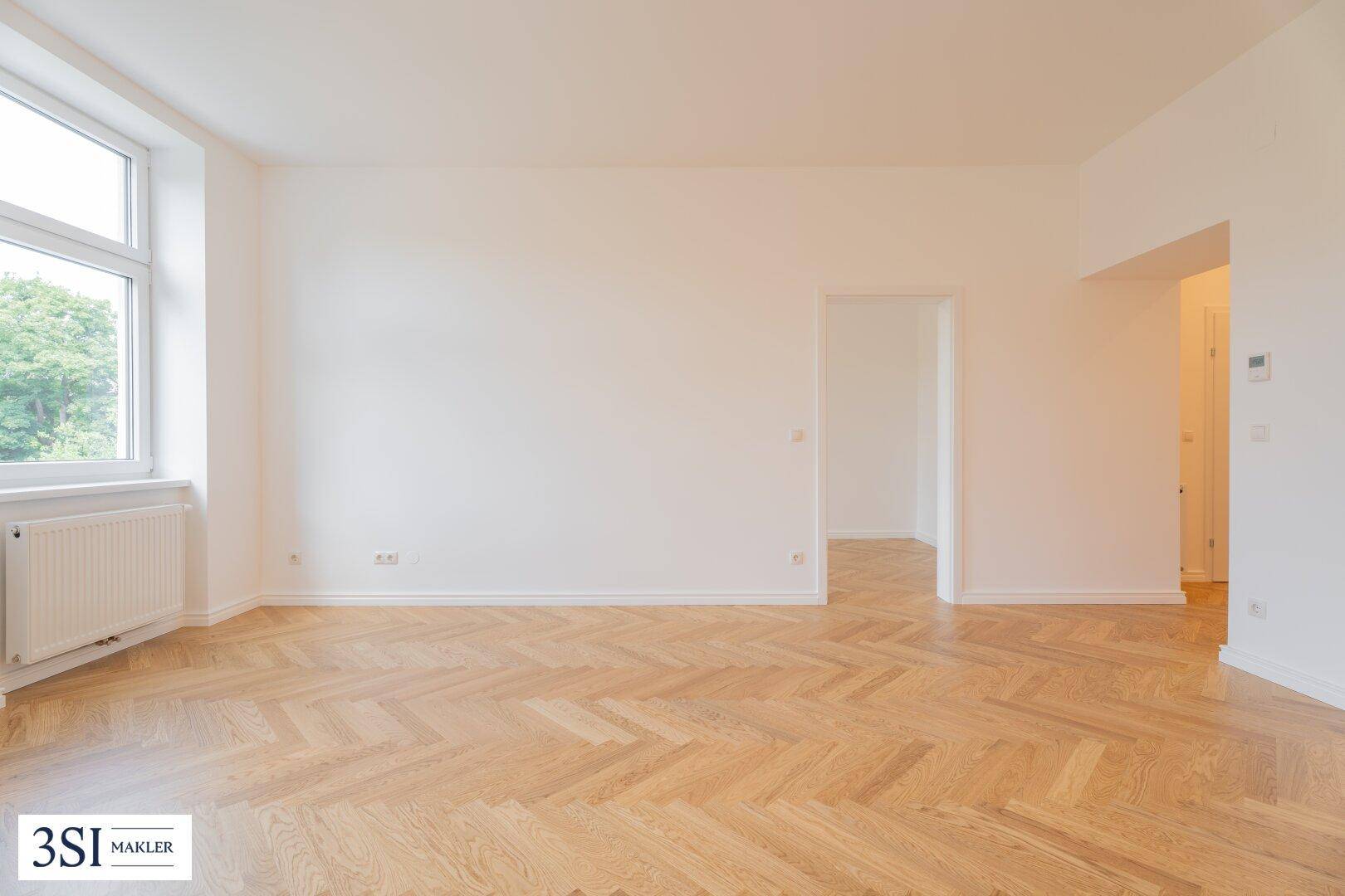 Heller, leerer Raum mit Fischgrätparkett und einem Fenster, das viel Tageslicht hereinlässt.