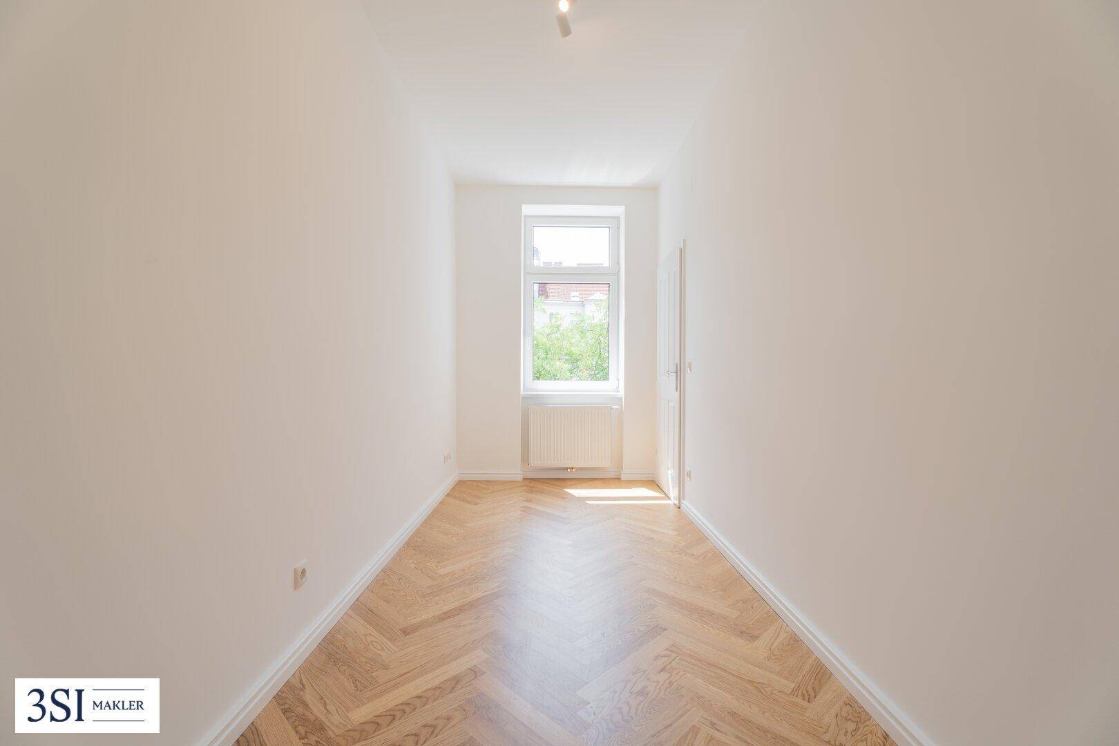 Schmaler, heller Raum mit Fischgrätparkett und einem Fenster, ideal als Schlaf- oder Arbeitszimmer.