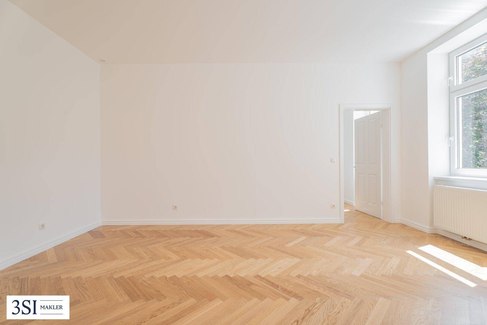 Heller, leerer Raum mit Fischgrätparkett und einem Fenster, das viel Tageslicht hereinlässt.