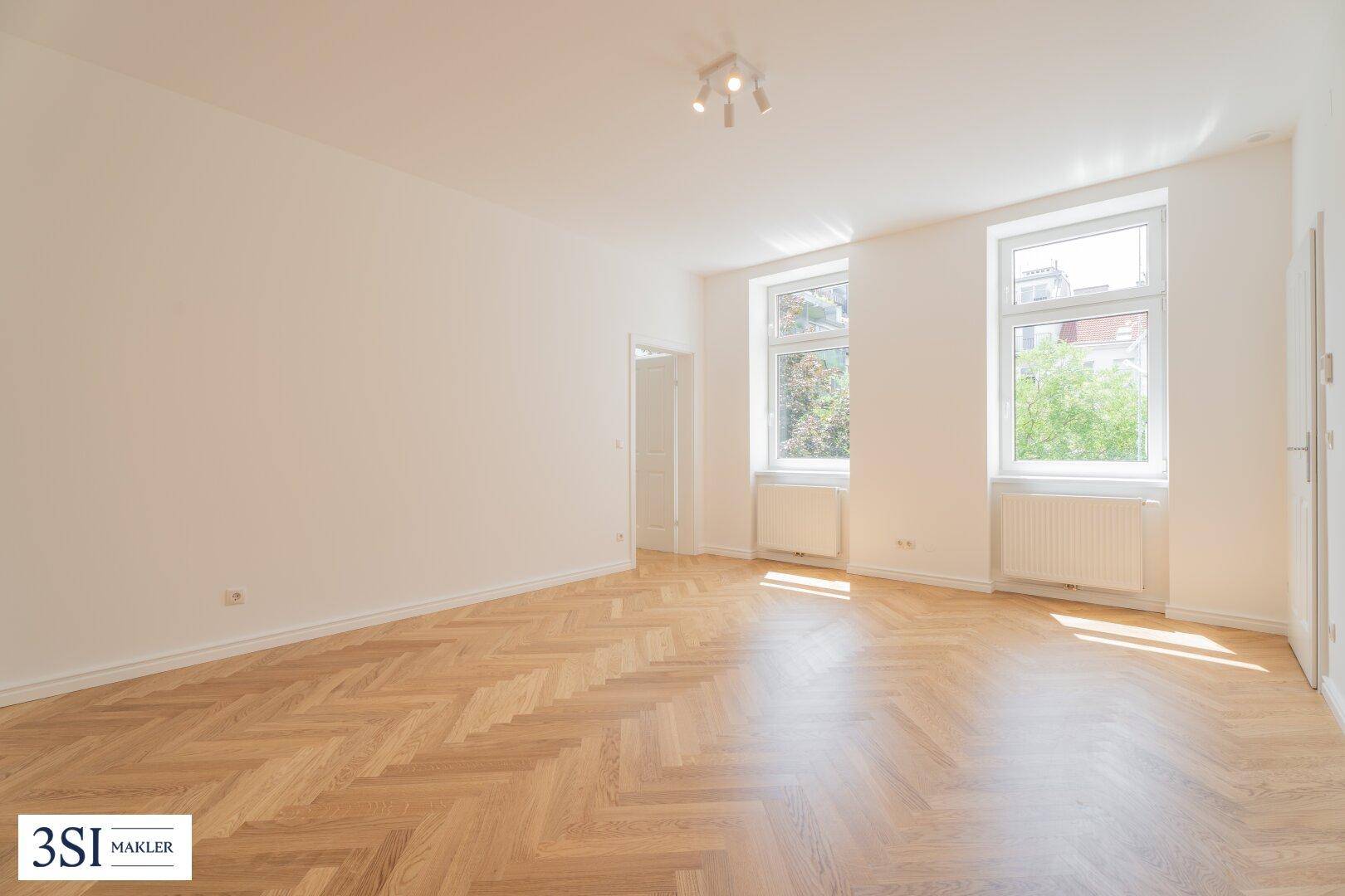 Geräumiges, leeres Wohnzimmer mit hellem Fischgrätparkett und großen Fenstern.