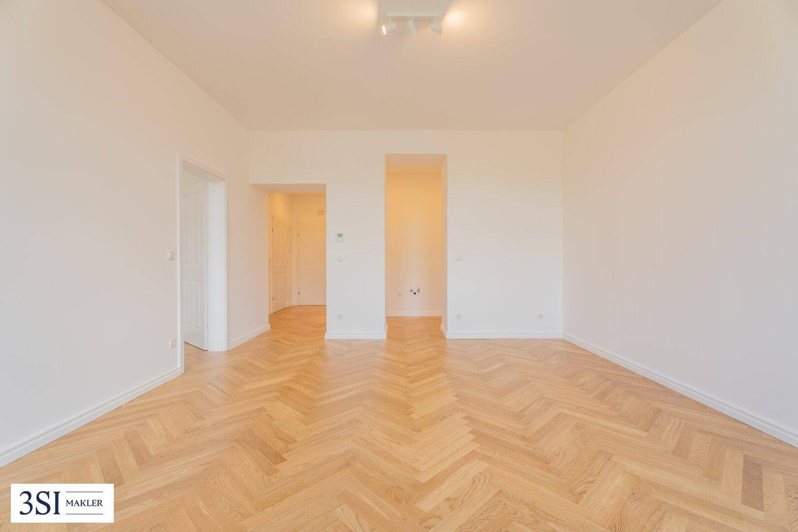 Geräumiges, leeres Zimmer mit hellem Fischgrätparkett und zwei Türöffnungen, die zu weiteren Räumen führen.
