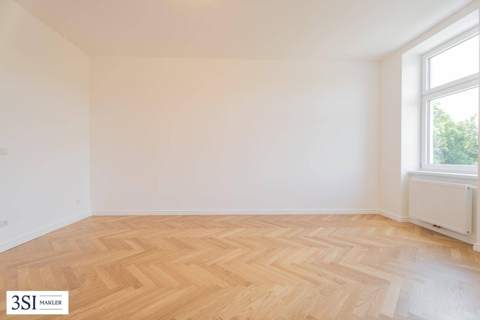 Geräumiges, leeres Zimmer mit hellem Fischgrätparkett und einem Fenster, das viel Tageslicht hereinlässt.