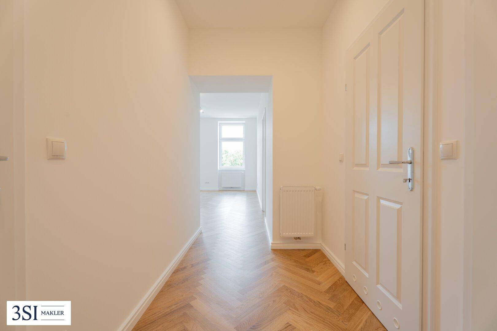 Langer, heller Flur mit Fischgrätparkett, der zu einem lichtdurchfluteten Zimmer mit Fenster führt.