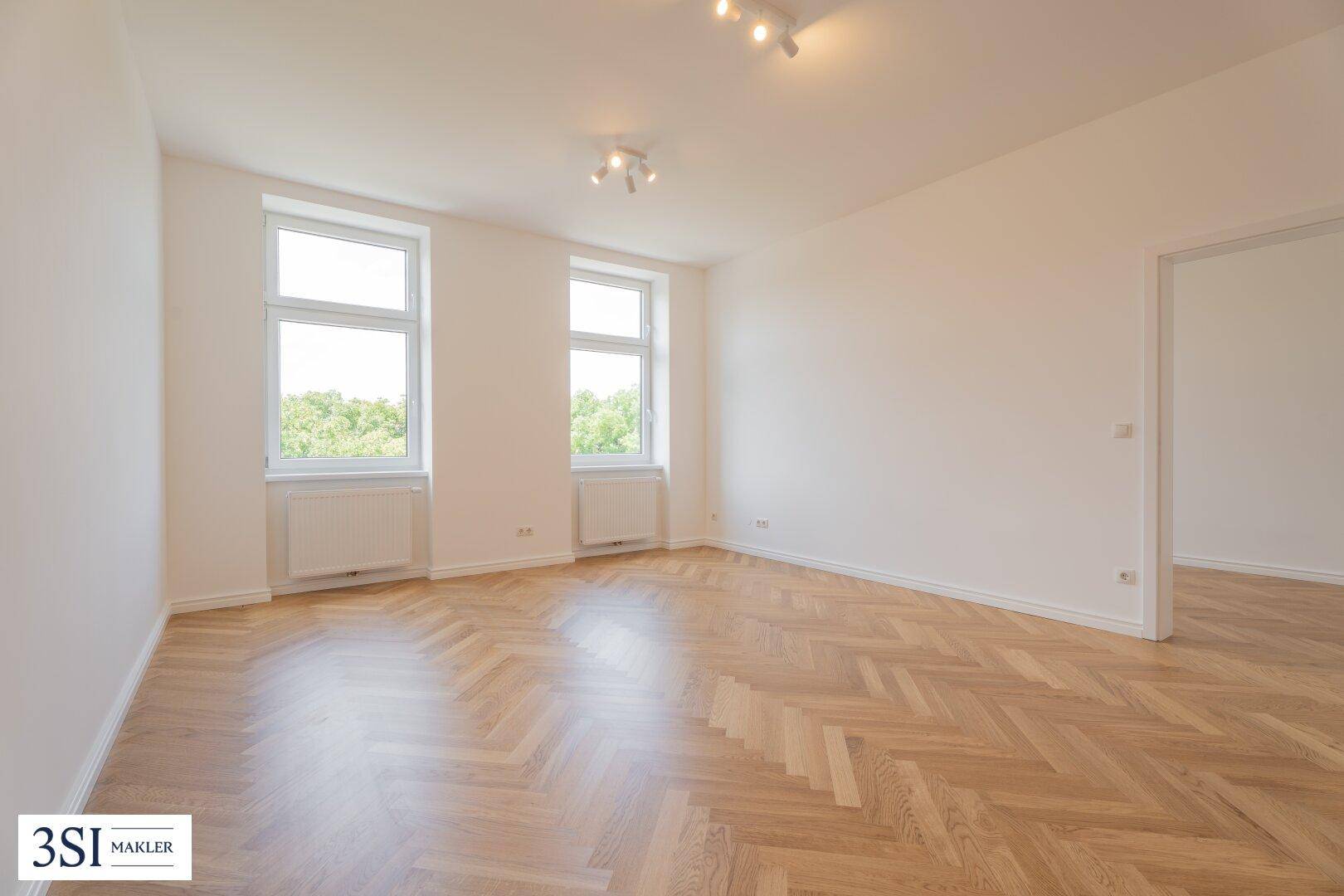 Helles, leeres Zimmer mit Fischgrätparkett und zwei großen Fenstern, bereit zur individuellen Gestaltung.