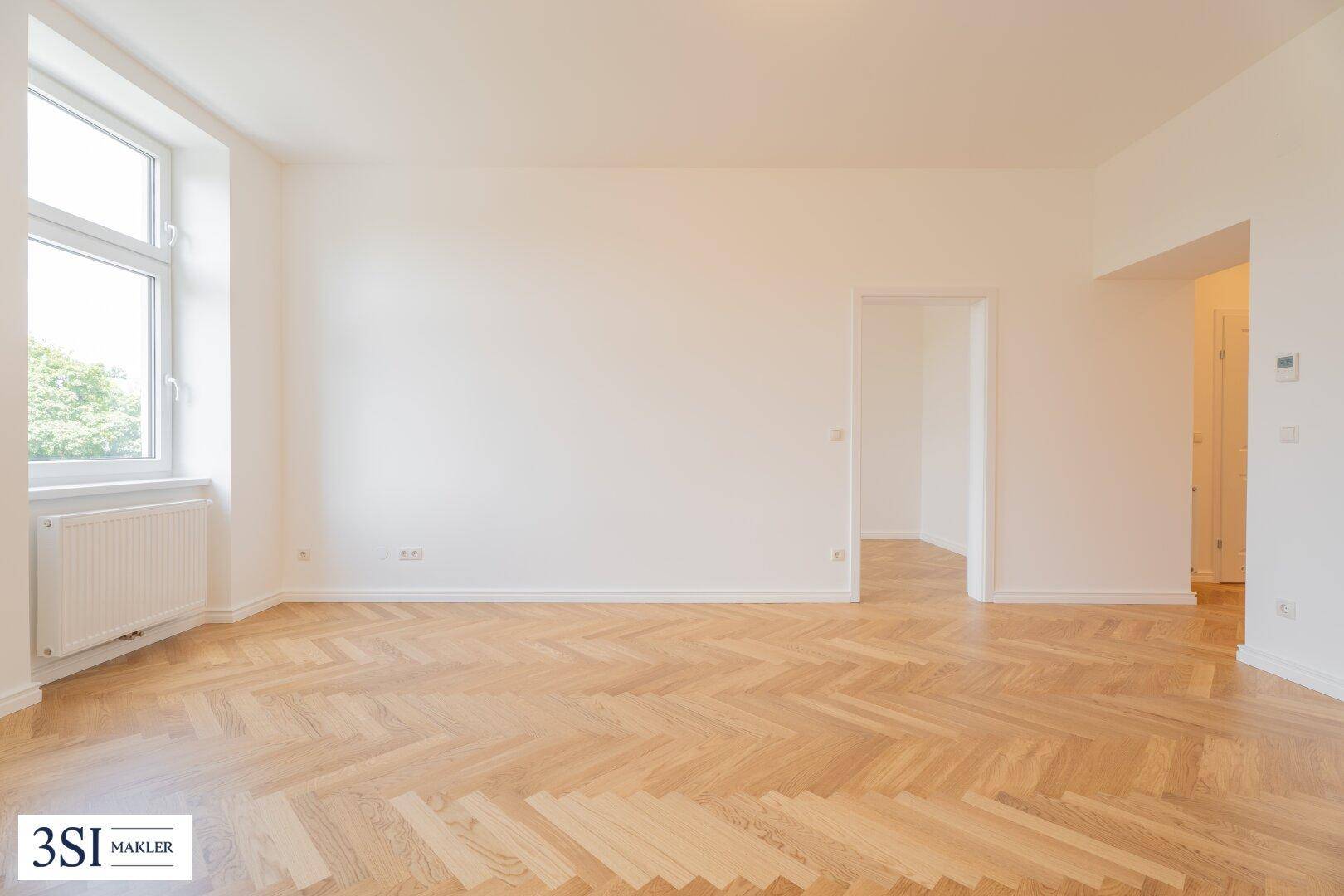Helles, leeres Zimmer mit Fischgrätparkett und einem Fenster, das den Blick ins Grüne freigibt.