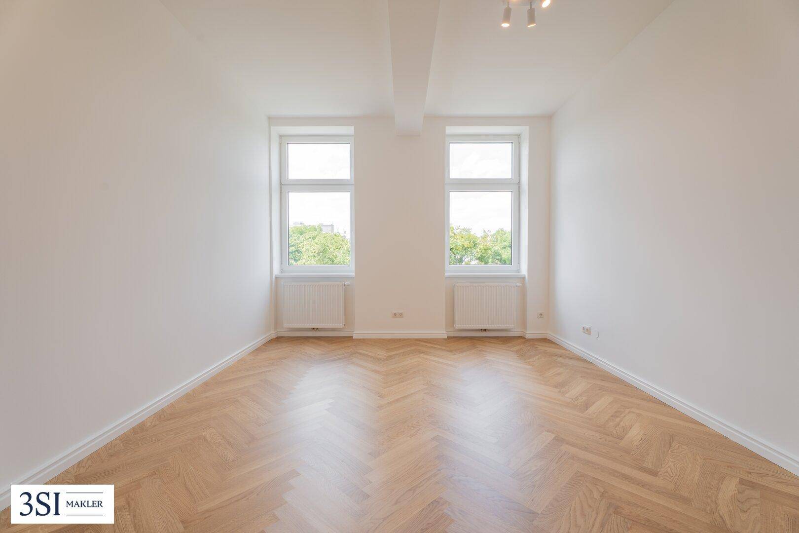 Geräumiges Zimmer mit Fischgrätparkett, hellen Wänden und zwei großen Fenstern.