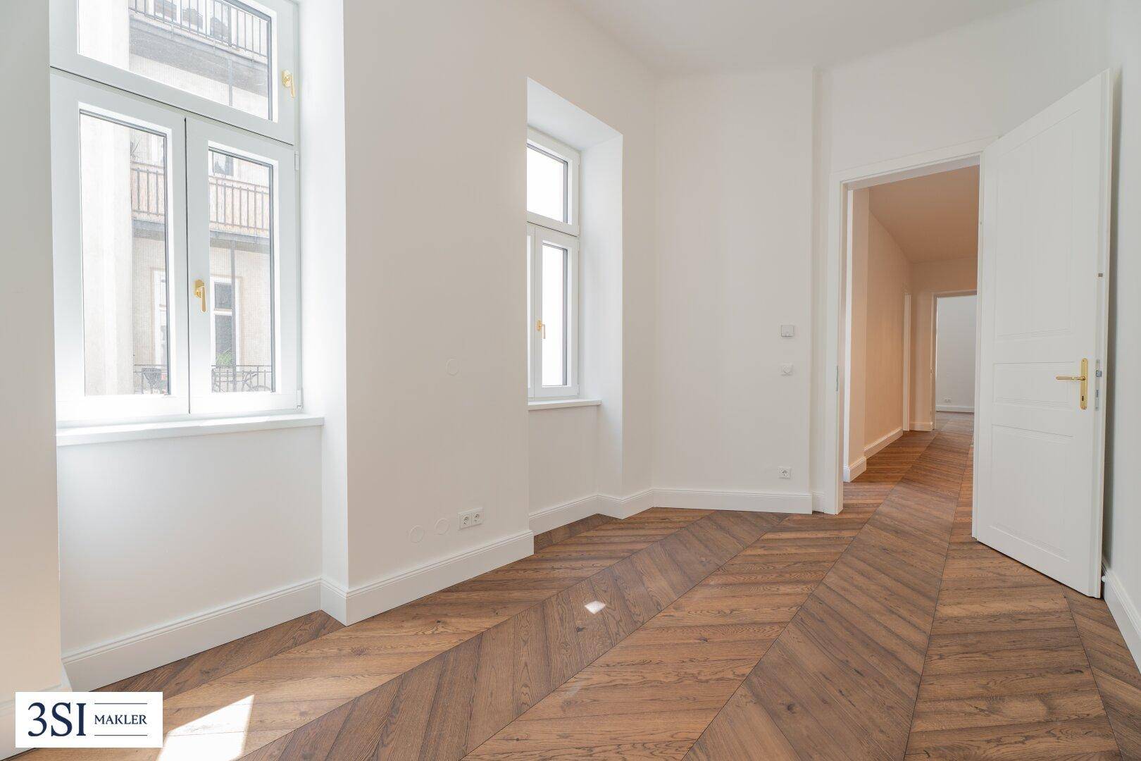 Heller Raum mit Fischgrätparkettboden und Fenstern, die auf einen französischen Balkon blicken.