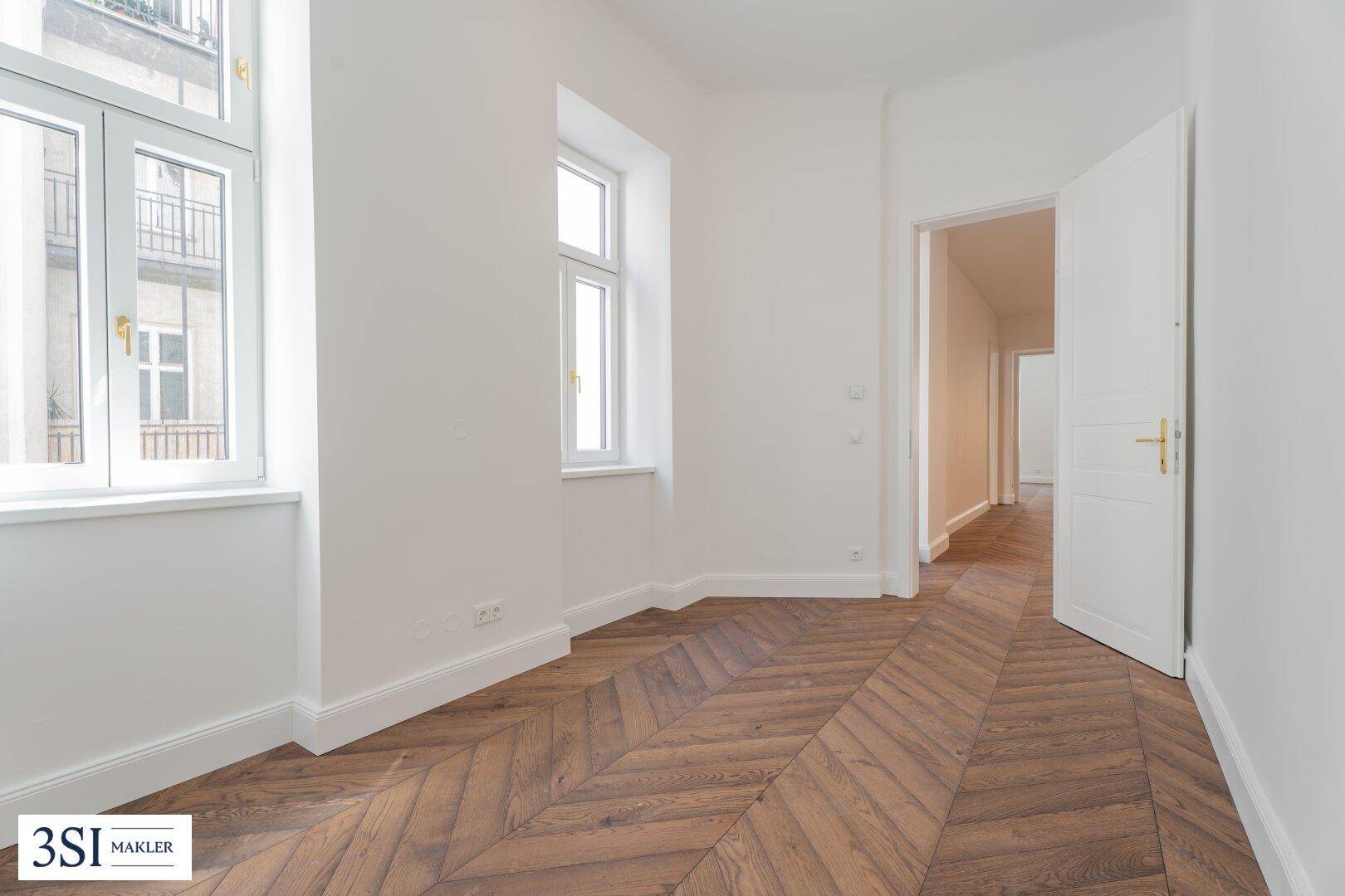 Freundliches Zimmer mit Fischgrätparkett und zwei Fenstern, bietet Zugang zu einem Flur.
