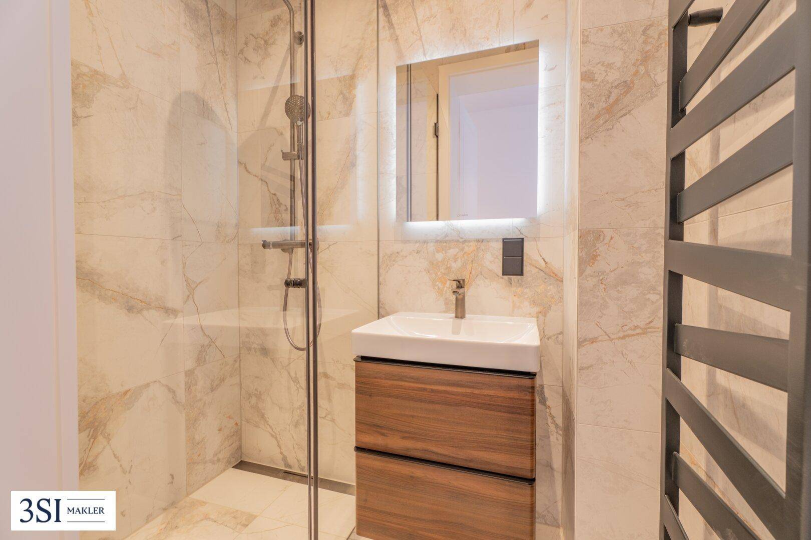 Helles Badezimmer mit Einzelwaschbecken, beleuchtetem Spiegel und begehbarer Dusche sowie Handtuchheizkörper.