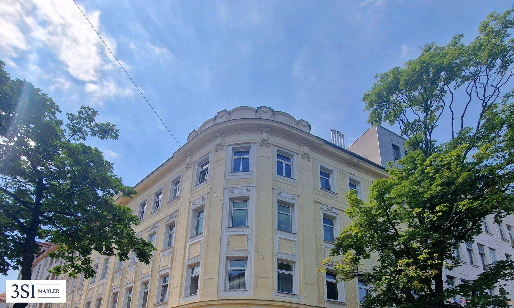 Historisches Eckgebäude mit klassischer Architektur, vielen Fenstern und umgeben von grünen Bäumen unter blauem Himmel.