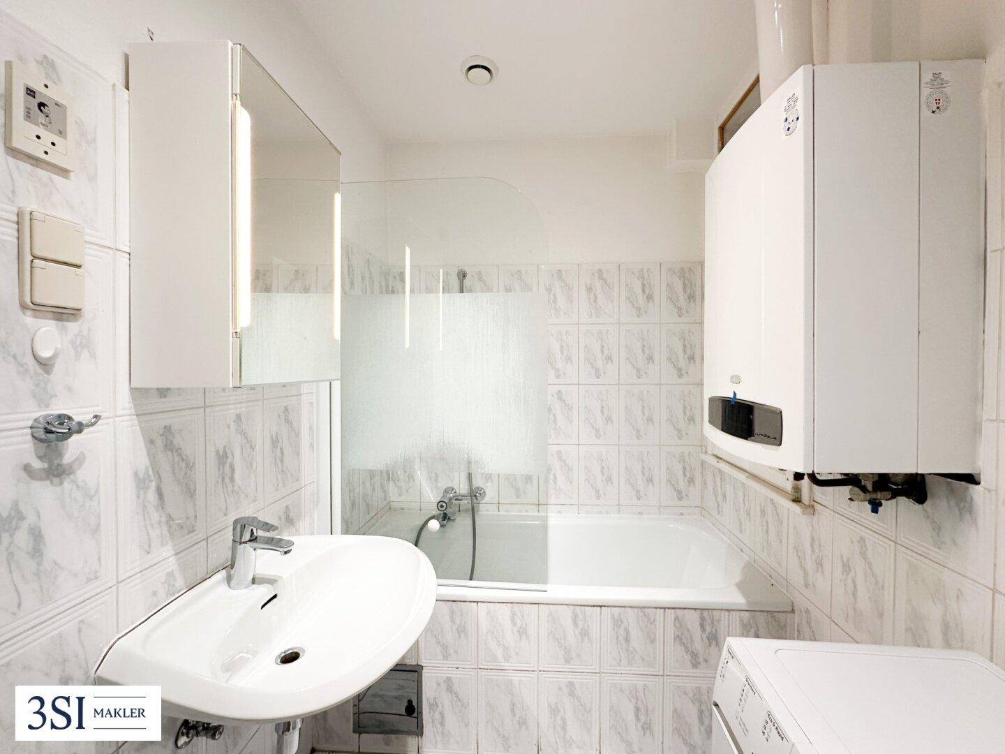 Badezimmer mit Badewanne, Waschbecken und Wandfliesen in hellem Muster, inklusive Warmwasserbereiter.