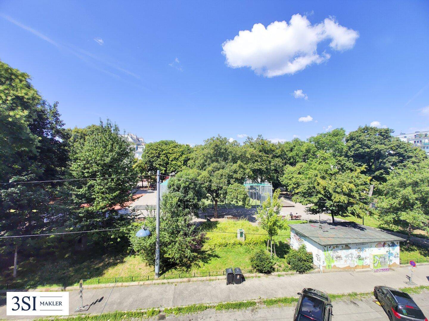 Weitläufiger Blick auf einen grünen Park mit Bäumen und einem Sportplatz unter blauem Himmel.