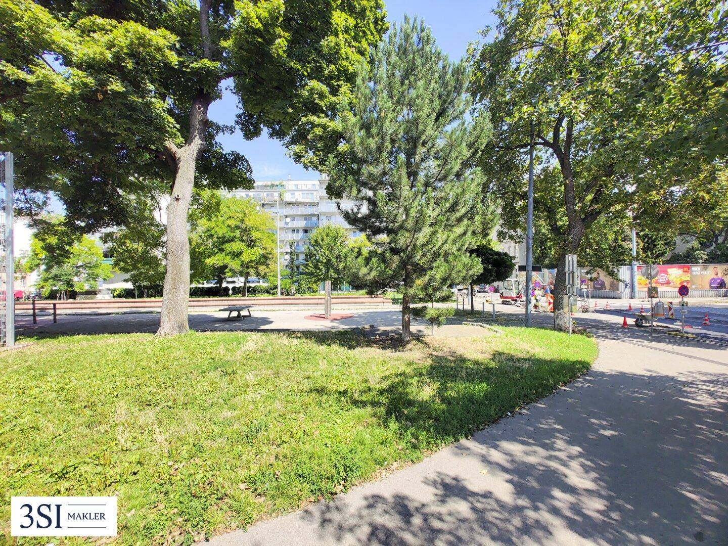 Grüne Parkanlage mit Bäumen und einem Gehweg, umgeben von städtischer Bebauung im Hintergrund.