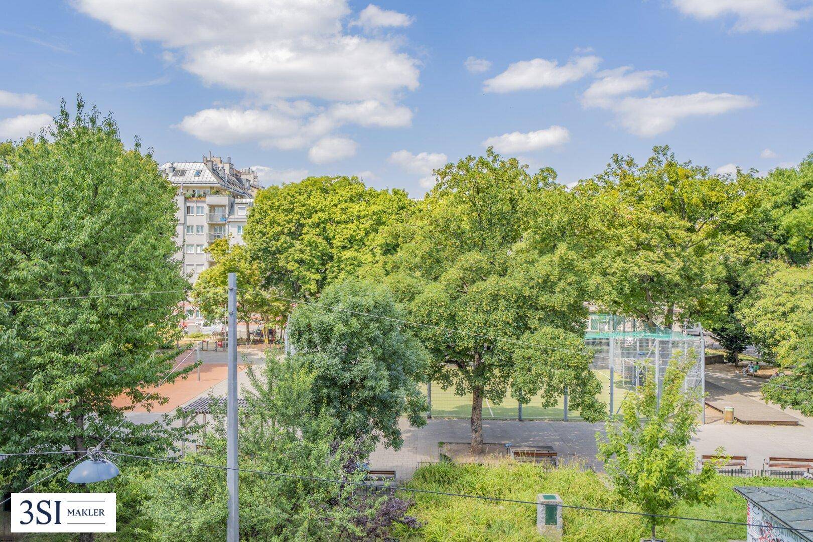 Weitläufiger Blick auf eine grüne Parkanlage mit Bäumen und einem Sportplatz unter blauem Himmel.