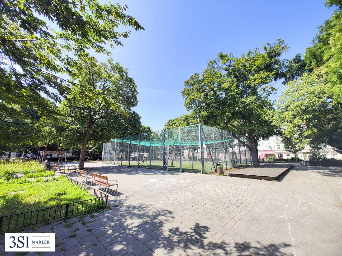 Öffentlicher Sportplatz mit Zaun und Sitzbänken, umgeben von Bäumen und Grünflächen in einem Park.