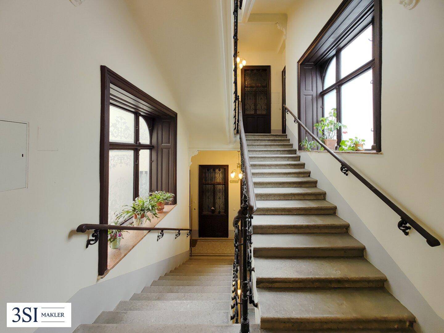 Helles Treppenhaus in einem Altbau mit großen Fenstern, Steinstufen und schmiedeeisernem Geländer.