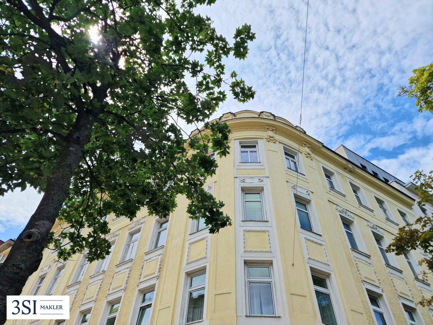 Eckgebäude eines Altbaus mit gelber Fassade und grünen Bäumen unter bewölktem Himmel.