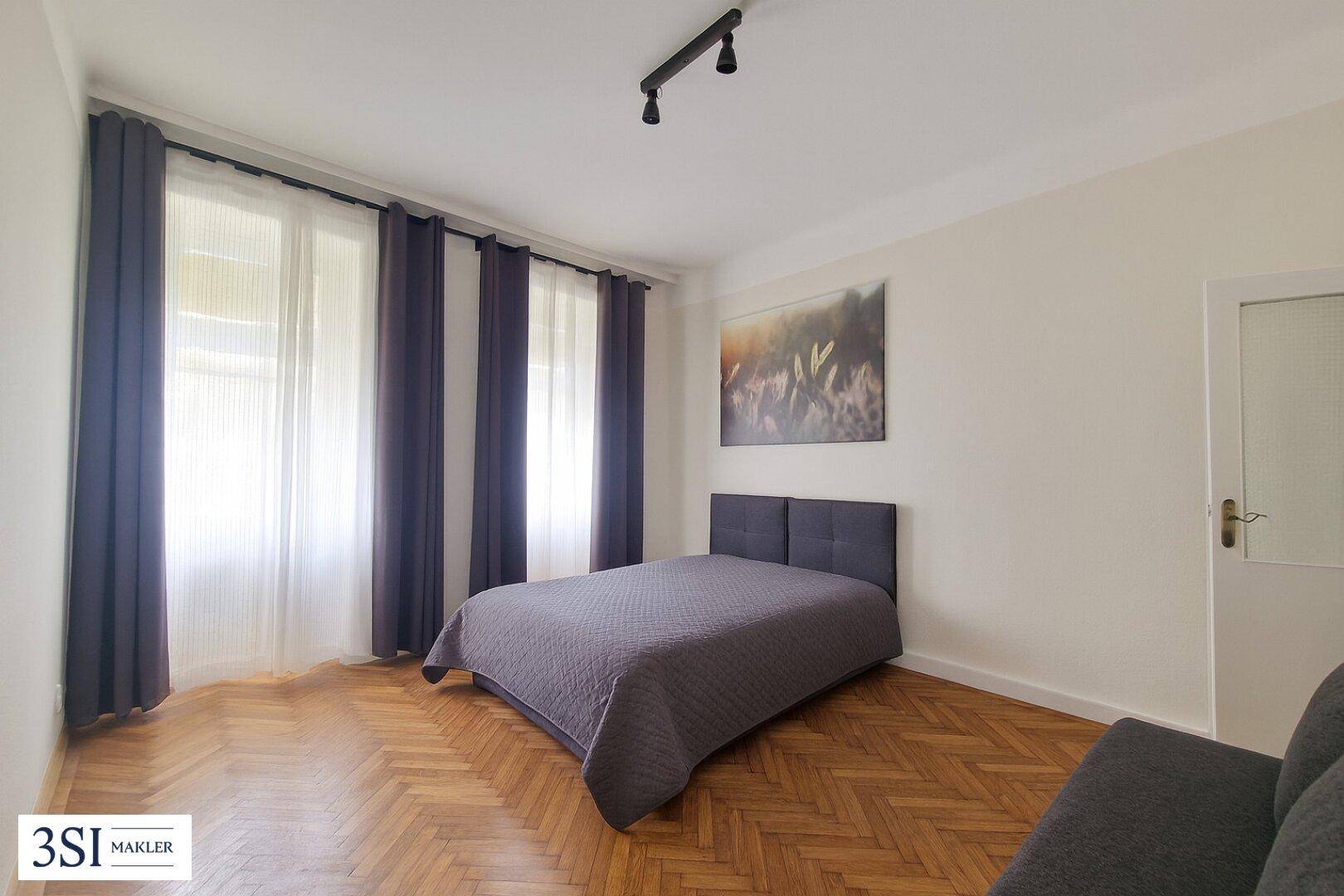 Gemütliches Schlafzimmer mit Doppelbett, Holzboden und großen Fenstern, die viel Tageslicht hereinlassen.