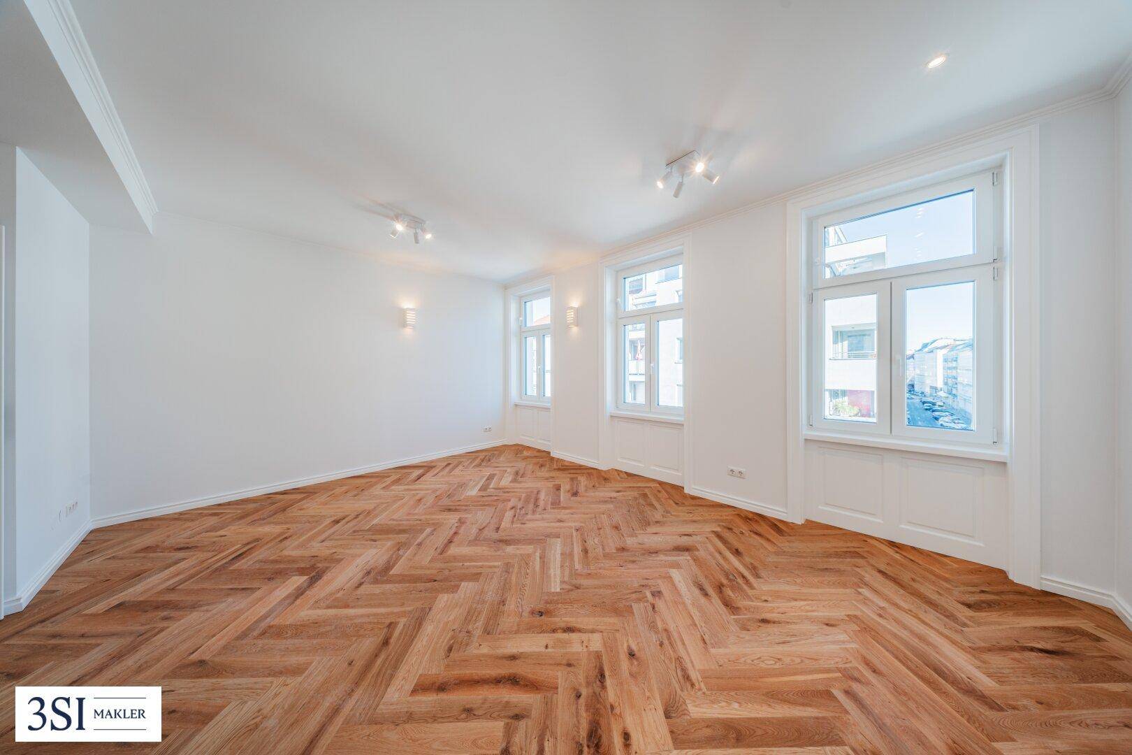Lichtdurchfluteter Raum mit elegantem Fischgrätparkett und mehreren großen Fenstern.