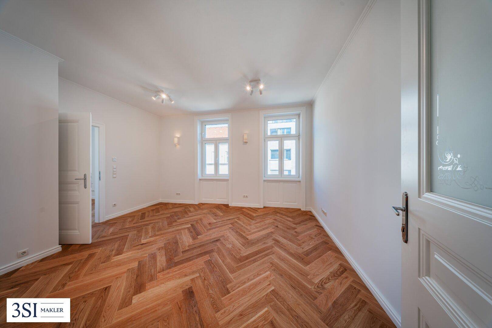 Geräumiges Zimmer mit Fischgrätparkett und zwei Fenstern, die viel Tageslicht hereinlassen.
