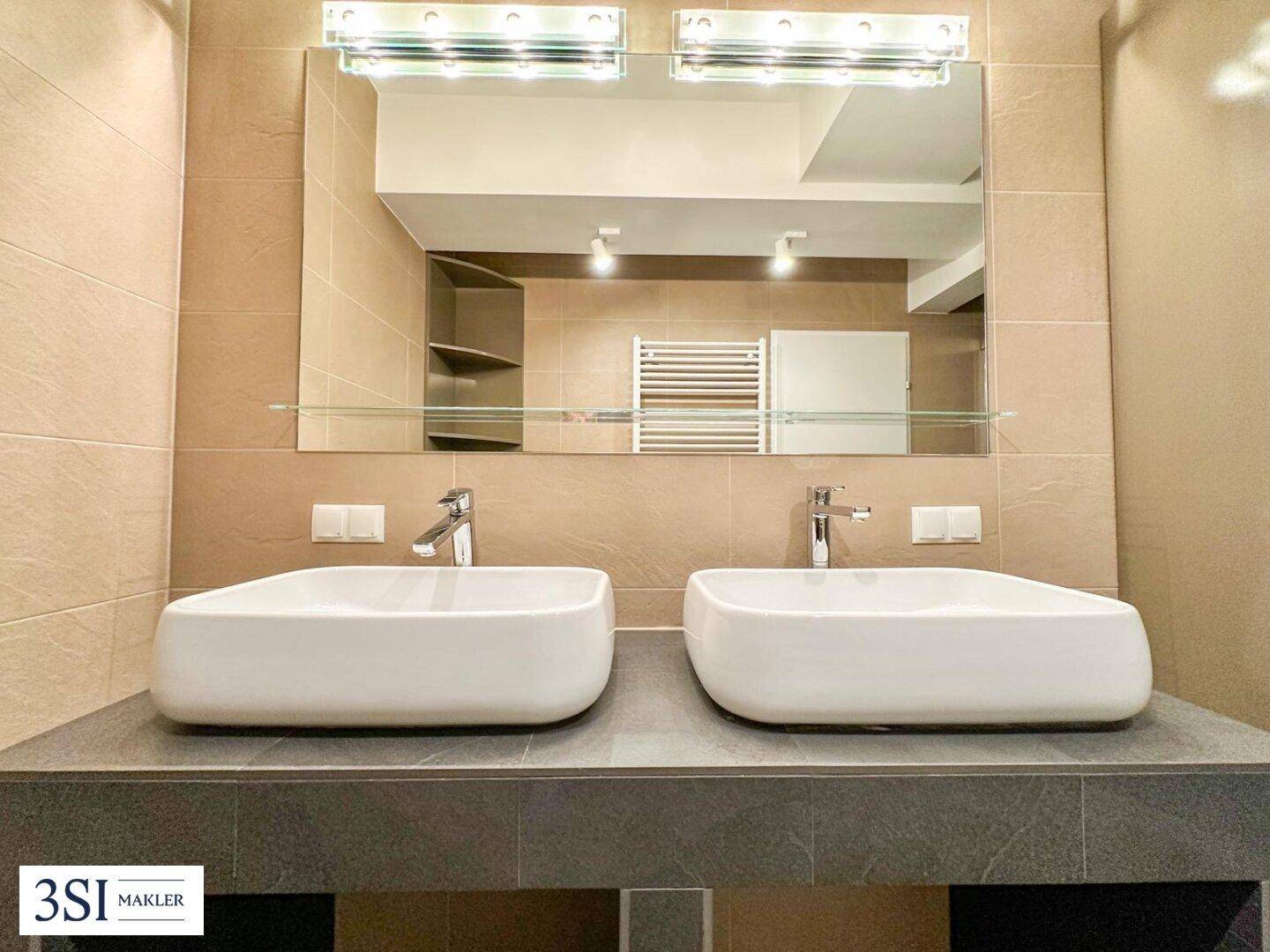 Badezimmer mit Doppelwaschbecken, großem Spiegel und beigefarbenen Wandfliesen.
