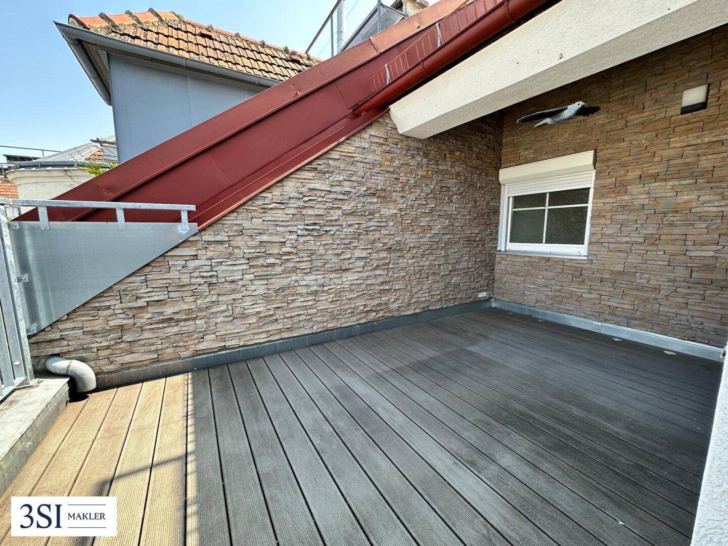 Private Terrasse mit Holzdeck, Steinwand und einem Fenster mit Rollladen.