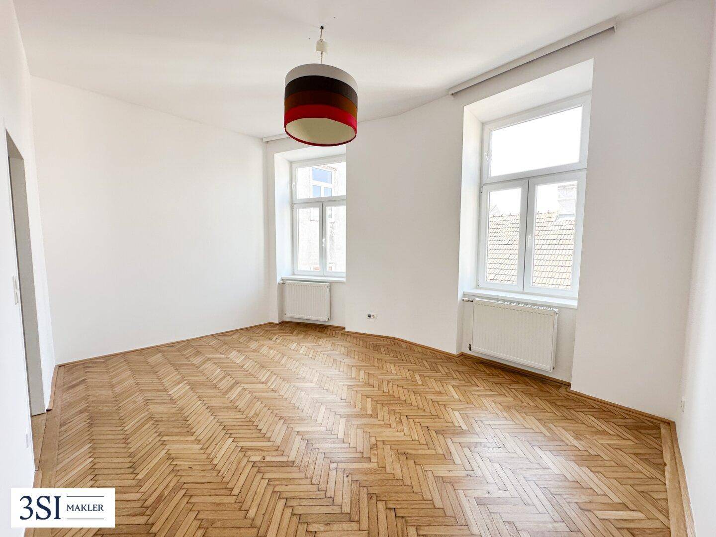 Helles Zimmer mit Fischgrätparkett, hohen Fenstern und einer modernen Deckenleuchte.