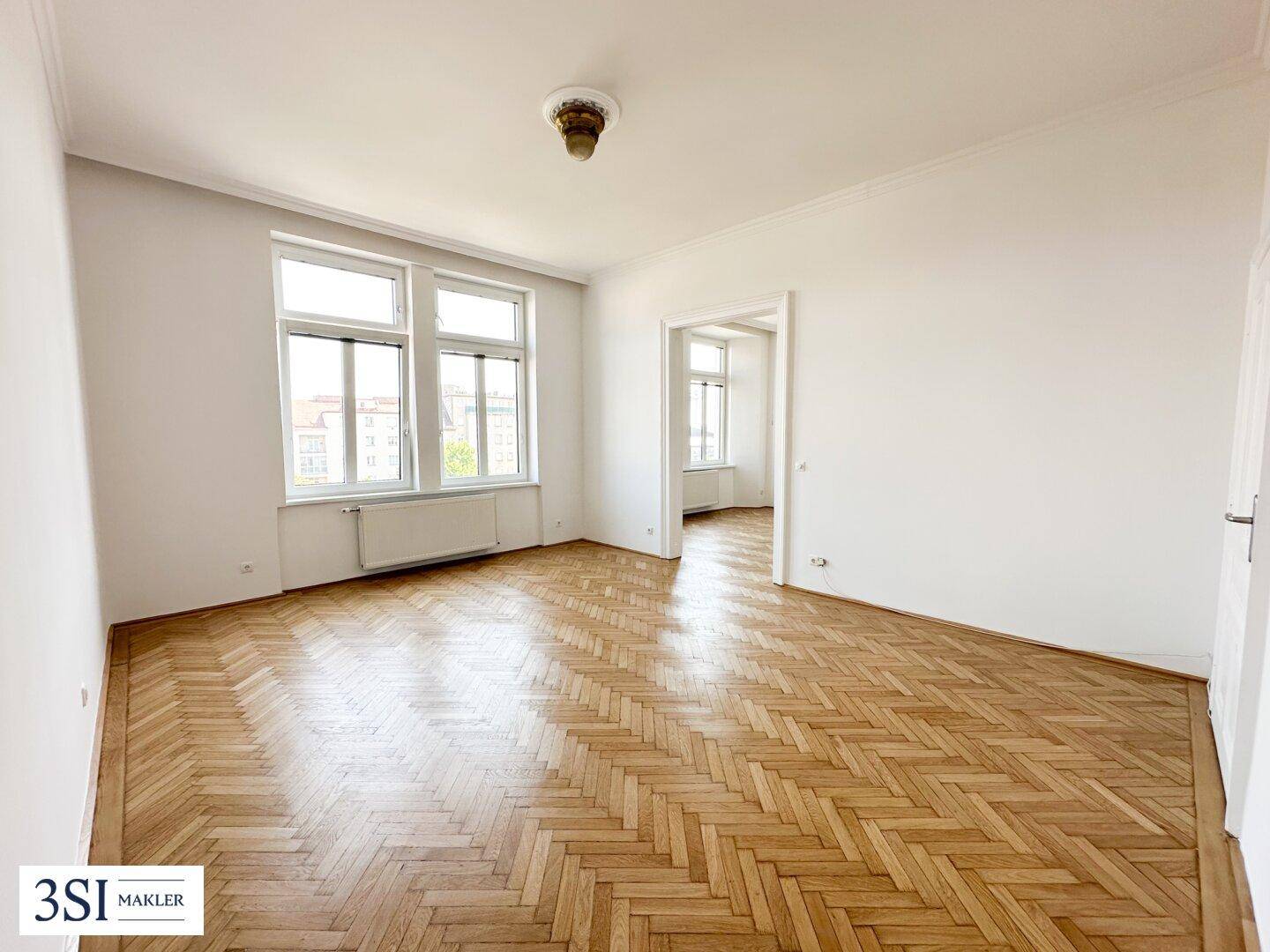 Helles Wohnzimmer mit Fischgrätparkett, hohen Fenstern und Zugang zu einem weiteren Raum.