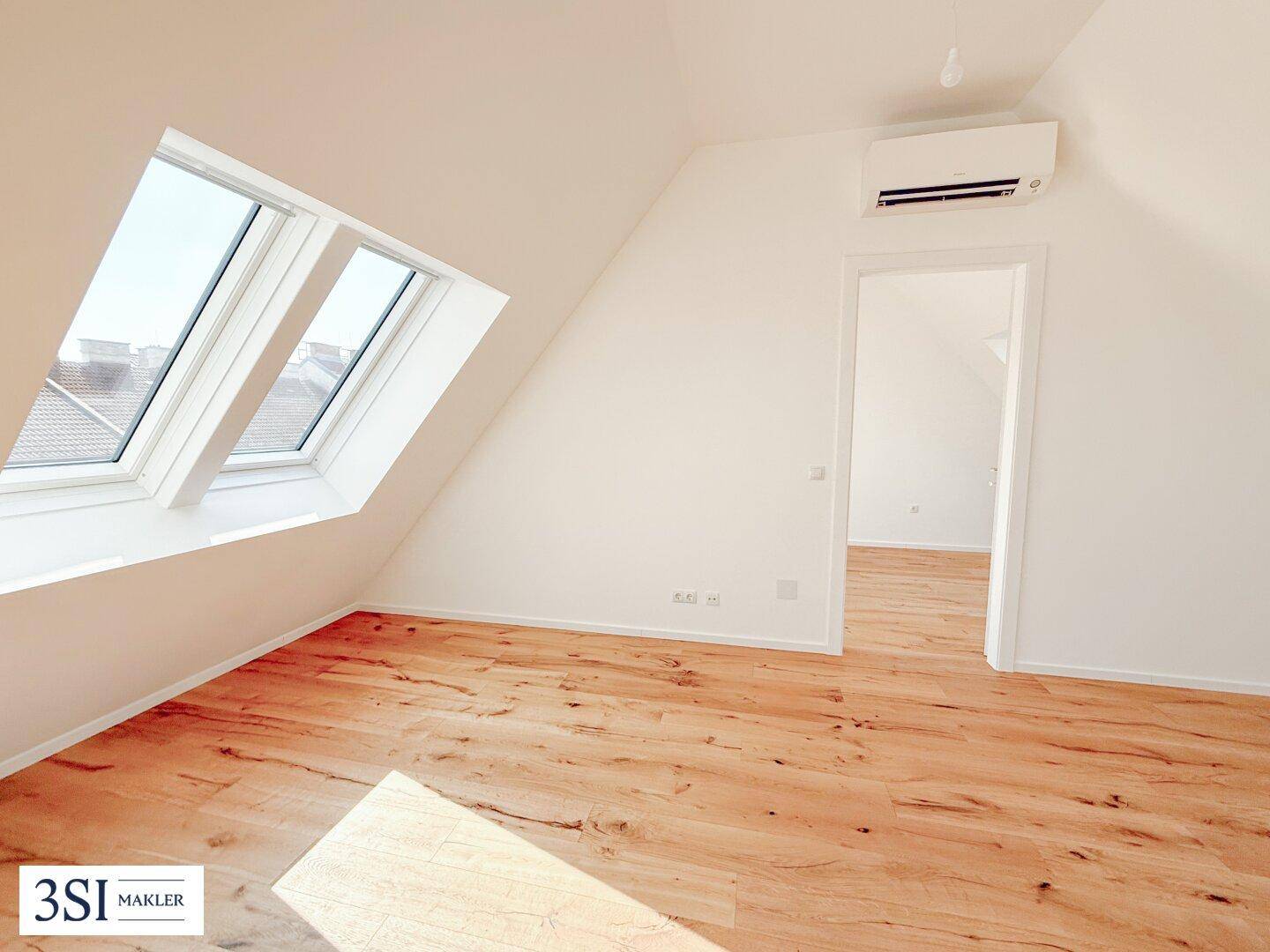 Heller Raum mit Holzboden, Dachschrägen, Dachfenstern und Klimaanlage, offen zu einem weiteren Zimmer.